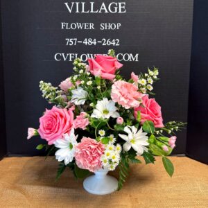 Pink and white floral arrangement in a white pedestal vase, featuring pink roses, white daisies, and carnations against a dark sign advertising Churchland's Village Flower Shop.