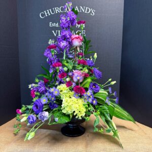 Tall, colorful floral arrangement in a black pedestal vase against a dark backdrop, featuring purple, pink, and blue blooms.",